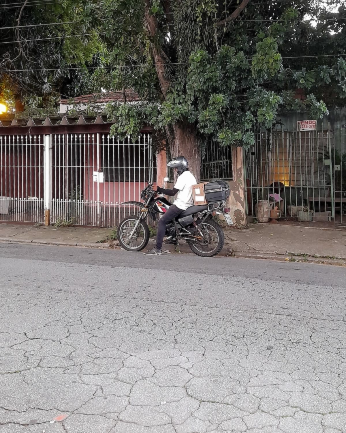 Motoboy com baú de entrega parado numa rua arborizada